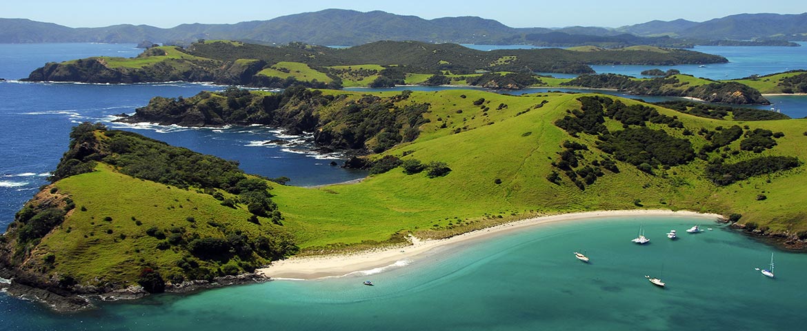 Wyprawy Nowa Zelandia 15 Odkrywanie Aotearoa. Zatoka Bay of Islands z zielonymi wzgórzami na wyspach w zatoce i piaszczystymi plażami. W zatoczkach wysp zakotwiczone małe jachty. Bezchmurne, niebieskie niebo. Rajski widok.